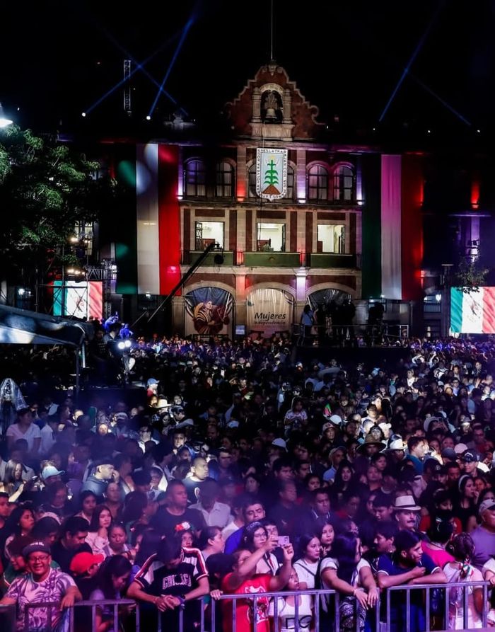 Miles de asistentes en un evento público nocturno frente a un edificio histórico iluminado con los colores de México